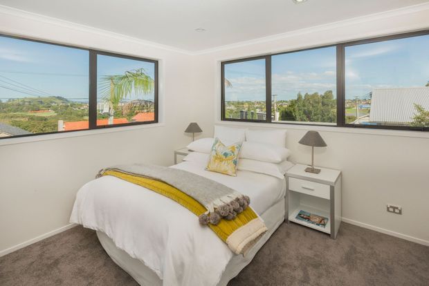 A bright, compact bedroom with a white upholstered bed dressed in white bedding, a grey throw with a mustard-yellow stripe, and a colourful accent cushion. Two white bedside tables with dark lamps flank the bed. Large black-framed corner windows provide expansive views of the surrounding landscape, while grey carpet, white walls, and simple cornices create a clean, contemporary interior.