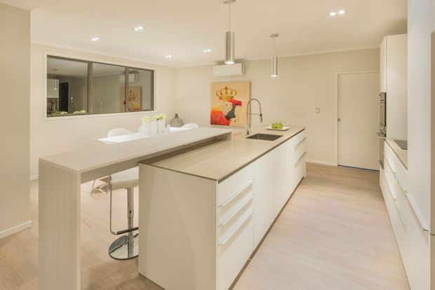 Bright, spacious contemporary kitchen with a large central island, integrated dining-height extension, white cabinetry, pale stone benchtops, stainless-steel sink and tapware, pendant lights, and light timber flooring. A small artwork and dining area add warmth to the otherwise clean, neutral interior.