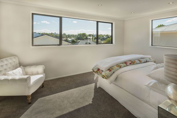 Bright, spacious bedroom with white upholstered furniture, a large bed layered with floral bedding, a tufted armchair, beige carpet, and broad black-framed windows overlooking a suburban neighbourhood. Recessed ceiling lights and restrained finishes create a clean, comfortable interior.