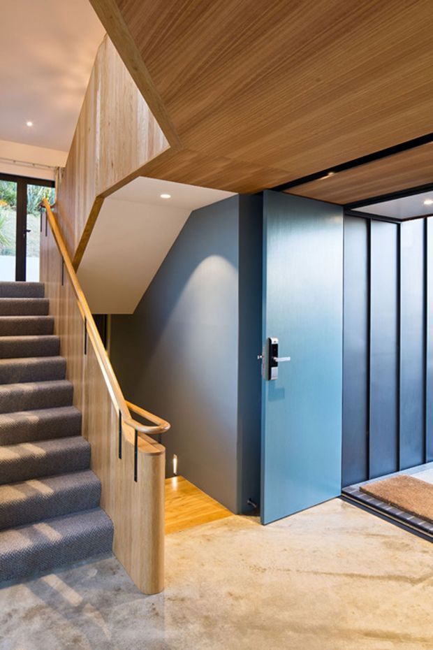 Contemporary residential entry hall featuring a carpeted staircase with timber balustrade and handrail, dark blue-grey walls, a blue pivot door, timber-lined ceiling, and polished concrete flooring. Full-height glazed panels bring light into the entrance, while warm timber detailing contrasts with the darker finishes.