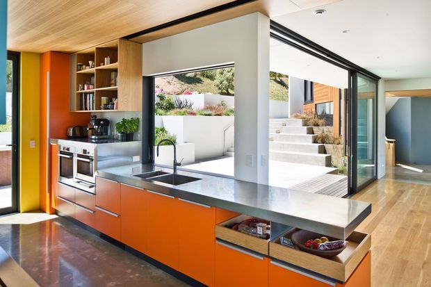 A bright, contemporary open-plan kitchen with vivid orange built-in cabinetry, stainless-steel appliances, a dark grey stone benchtop, timber ceiling, and integrated open shelving. Large glazed sliding doors connect the kitchen to a landscaped courtyard and exterior steps, while timber flooring continues through the adjacent circulation space.