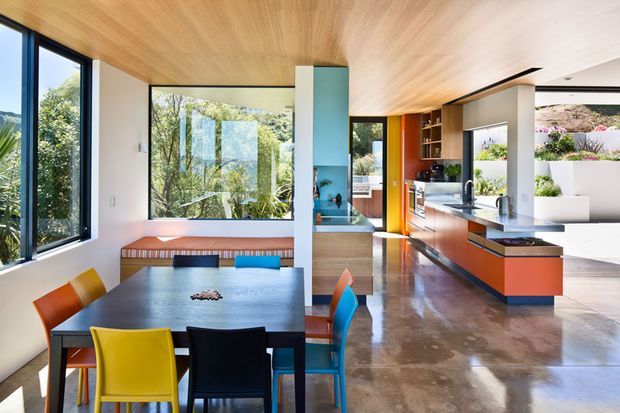 Bright open-plan contemporary interior combining a dining area and kitchen. The room features full-height glazing, a warm timber-lined ceiling, polished concrete flooring, white walls, and a bold orange and dark-toned kitchen with an island. Colourful dining chairs and built-in bench seating add playful accents, while garden landscaping is visible through the windows.