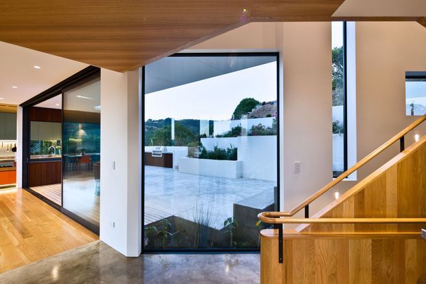 Contemporary interior view of a split-level entry and stairway, featuring extensive floor-to-ceiling glazing, warm timber ceiling and stair balustrade, polished concrete flooring, and views toward a landscaped terrace and pool-like courtyard outside. A kitchen or living area is visible through the glazed opening on the left.