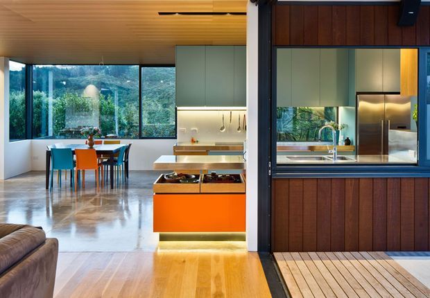 Open-plan contemporary kitchen and dining area with a vivid orange island, timber cabinetry and ceiling, polished concrete flooring, and large glazed windows overlooking a lush wooded landscape. A dark-framed pass-through window connects the kitchen to an exterior timber deck or adjacent outdoor area.
