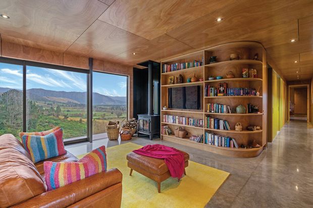 Warm, timber-lined living room with a large built-in open bookcase, leather sofa and ottoman, colourful woven cushions, a yellow area rug, polished concrete flooring, and expansive corner glazing overlooking a rural mountain landscape. Recessed ceiling lighting and a hallway connection are visible.