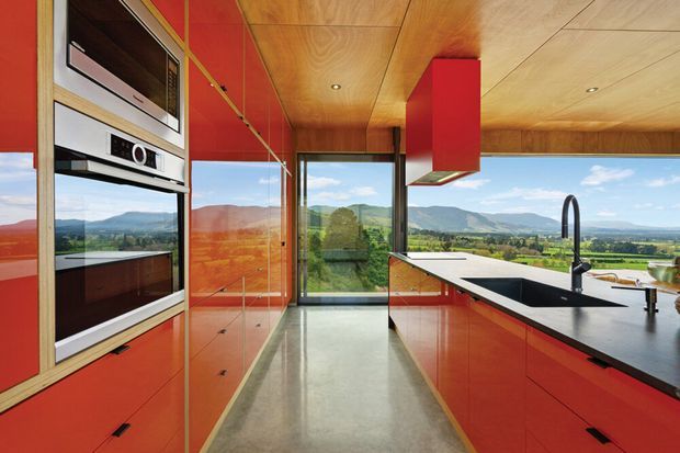 A bold contemporary kitchen with glossy red cabinetry, integrated stainless-steel appliances, a dark island benchtop with sink and curved black tap, and a warm timber-lined ceiling. Full-height glazing opens to expansive green rural hills and mountain views, while reflective surfaces and the polished floor enhance the bright, dramatic interior.