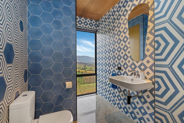 A compact, highly patterned bathroom with blue-and-white geometric wall tiles, including hexagonal and diamond motifs. The room contains a wall-mounted white basin, toilet, arched mirror, black-framed full-height window, and a grey polished floor beneath a warm timber ceiling.