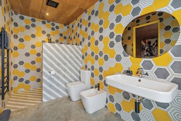 A bold, retro-inspired bathroom featuring vivid yellow walls covered in geometric hexagonal and octagonal patterned tiles in grey, white and beige. The room includes a white pedestal-style basin with exposed chrome plumbing, matching toilet and bidet, a glass shower enclosure with diagonally striped grey-and-white tiles, a large circular mirror, and a warm timber-panelled ceiling.