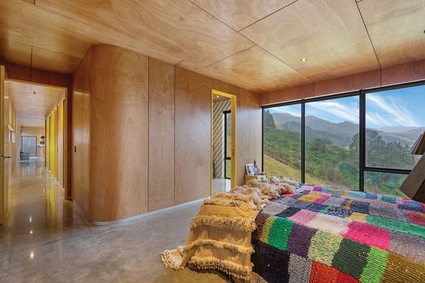 A warm, wood-lined bedroom with built-in timber cabinetry and a polished concrete floor. A colourful woven bedspread and textured throw sit on the bed in the foreground, while large floor-to-ceiling windows provide expansive views of a green mountainous landscape. A timber-lined hallway and additional rooms extend beyond the bedroom.