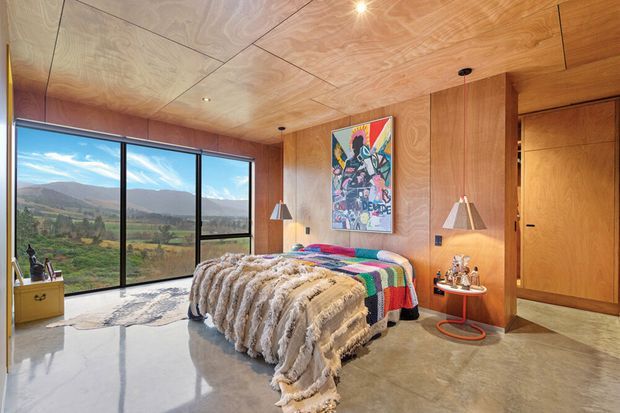 A warm, wood-lined bedroom with a large bed dressed in a multicoloured woven throw and textured blanket. Floor-to-ceiling glazing opens to a broad mountain and rural valley view, while integrated timber wall and ceiling panelling, bedside lighting, artwork, and a flush timber door create a cohesive contemporary retreat. The polished concrete floor adds contrast to the natural timber interior.