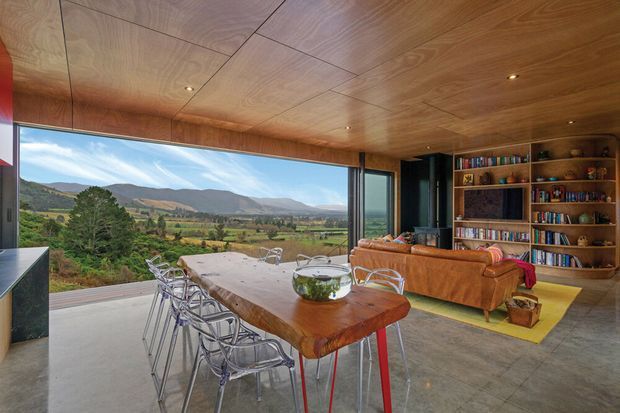 A warm, wood-lined open-plan living and dining space with a large timber dining table and metal-framed chairs beside expansive floor-to-ceiling glazing. A leather sofa, built-in timber bookcase, and recessed ceiling lighting create a relaxed contemporary interior, with broad rural mountain views beyond the glass.