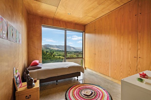 Warm wood-lined bedroom with built-in floor-to-ceiling wardrobes, a single upholstered bed, bedside furniture, a colourful circular rug, and a large window overlooking a rural valley and mountains. The room has timber wall and ceiling panelling and a polished concrete floor.