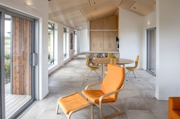Bright open-plan interior with a dining area and compact kitchen at the rear, furnished with mid-century-inspired timber and orange seating. Large glazed doors and windows line the left side, opening to a timber deck and landscape. The room has a pale timber-lined ceiling, white walls, large-format light grey stone or concrete-look flooring, and integrated timber cabinetry.