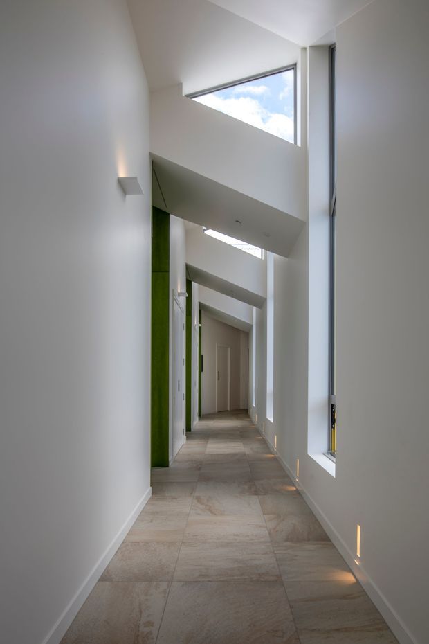 A long, narrow minimalist hallway with tall white walls, pale stone-look tiled flooring, high clerestory windows, and dramatic angular overhead openings. Slim vertical windows bring in daylight, while recessed wall lights and small floor-level uplights create a clean, gallery-like atmosphere. Green-painted vertical architectural elements add a subtle accent along the corridor.