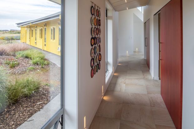A bright contemporary hallway with a long corridor, pale stone or concrete-look flooring, white walls, recessed ceiling lighting, and large floor-to-ceiling glazing. A vivid red timber door and colourful circular wall artwork provide accents. Through the glazing, a yellow single-storey wing and landscaped native-style planting are visible.