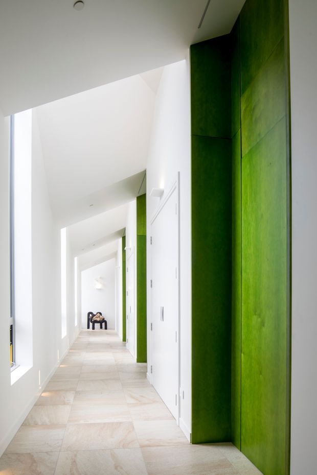 A bright, elongated interior hallway with white walls, ceiling and doors, pale large-format stone or porcelain floor tiles, and dramatic full-height vivid green built-in wall panels or cabinetry along the right side. Narrow vertical windows bring in abundant natural light. The corridor leads toward a small dark chair at the far end, creating strong linear perspective. The design is clean, minimalist and contemporary, with a high-contrast green feature surface against a predominantly white palette.