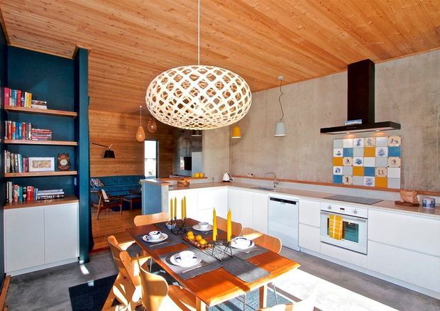 Open-plan kitchen and dining area with white built-in cabinetry, integrated appliances, a long island, and a timber dining table. The room features warm timber ceiling and wall panelling, dark blue built-in shelving and seating, a raw concrete feature wall, colourful tiled splashback, and a large woven pendant light.