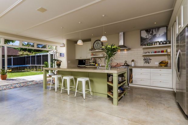Bright, open-plan kitchen with a large green island and three white stools, integrated white cabinetry, white subway-tile splashback, stainless-steel appliances, polished concrete flooring, pendant lighting, and a broad glazed opening to a backyard. The kitchen has a relaxed contemporary character with abundant natural light.