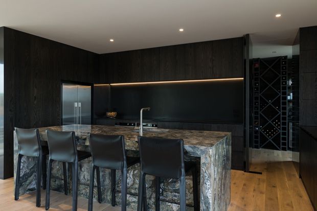A dramatic contemporary kitchen with dark timber cabinetry, a black backsplash, integrated appliances, and a large stone island with four black leather bar stools. Warm LED strip lighting highlights the cabinetry, while a timber floor and a glass-fronted wine storage area add warmth and functionality.