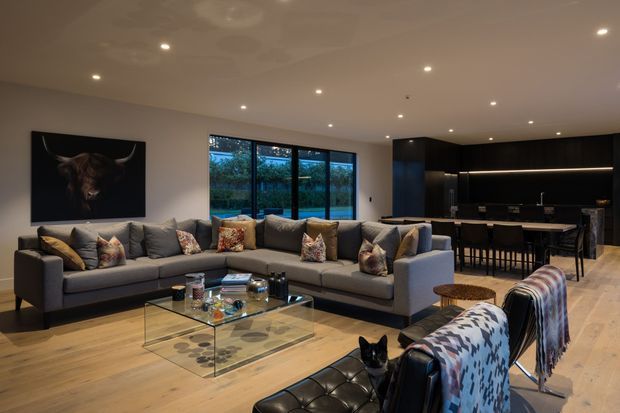 A spacious contemporary open-plan living room with a large L-shaped grey sectional sofa, black leather accent seating, a glass coffee table, and warm timber flooring. The room includes extensive dark built-in cabinetry and a bar or kitchen area at the rear, broad garden-facing windows, recessed ceiling lighting, and a dark feature artwork. The palette is warm and neutral with strong black accents.