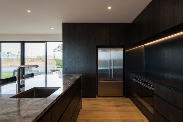 Contemporary dark-toned kitchen with extensive black-stained timber cabinetry, a large stone island with integrated sink and brushed metal tapware, stainless-steel appliances, timber flooring, and recessed linear lighting. Full-height glazing opens the kitchen toward an outdoor setting.