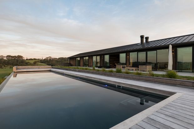 Wide exterior view of a long, low modern residence beside a large reflecting swimming pool. The house has a dark standing-seam metal pitched roof, extensive floor-to-ceiling glazing, dark cladding, and a broad timber deck with outdoor seating. The landscaped setting appears open and rural, with the pool and deck dominating the foreground.