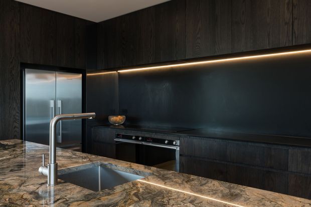 A dark, sophisticated contemporary kitchen featuring full-height charcoal timber cabinetry, integrated stainless-steel appliances, a black splashback, under-cabinet linear lighting, and a large natural-stone island with an undermount sink and brushed metal tap.