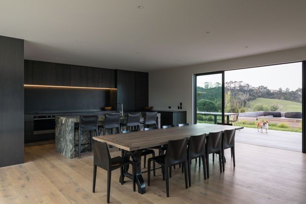 A contemporary open-plan kitchen and dining area with dark full-height cabinetry, a stone kitchen island, integrated warm strip lighting, and a long timber dining table with black chairs. Wide glazed sliding doors open to a covered outdoor deck and landscaped rural surroundings.