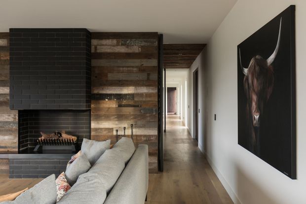 A contemporary living room and hallway with a dramatic black tiled fireplace, rustic reclaimed timber wall and ceiling lining, polished concrete hearth, and warm timber flooring. A large neutral sectional sofa occupies the foreground, while a long corridor extends toward the rear of the home. A monochrome bull artwork provides a bold accent on the white wall.