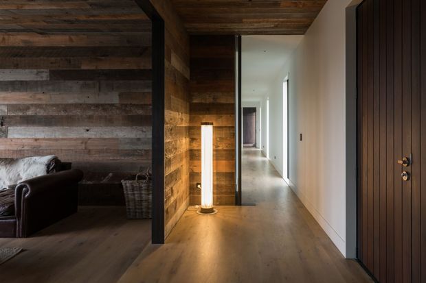 A warmly lit residential hallway with wide-plank timber flooring, reclaimed timber wall and ceiling cladding, and a dark timber entry door. The hallway opens toward a bright minimalist corridor, while a partially visible living room with a leather armchair sits to the left. A vertical floor lamp adds a soft glow beside the timber feature wall.