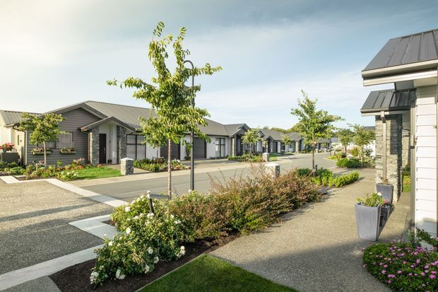 A landscaped residential development viewed from a pedestrian walkway, showing a row of compact single-storey homes with dark horizontal cladding, light trim, pitched metal roofs, covered entry porches, and paved driveways. Young trees, garden beds, flowering plants, and neat paths create a well-maintained suburban streetscape.