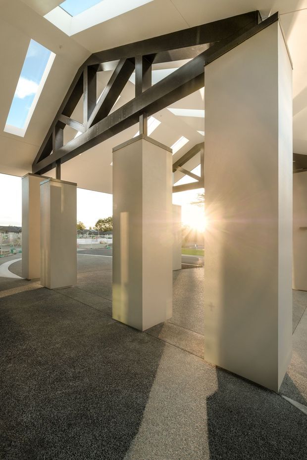 A dramatic architectural interior or covered public space with tall white rectangular columns, exposed dark steel beams, and multiple skylights. Strong low-angle sunlight enters through the open sides and roof glazing, casting long shadows across a dark textured floor. The space appears minimal, structural, and possibly transitional between interior and exterior.