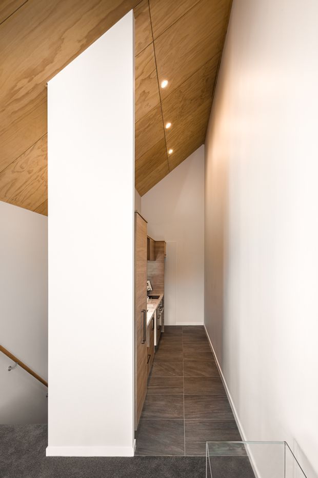 A narrow, elongated kitchen corridor beneath a steeply sloped timber-lined ceiling. The space features warm wood cabinetry along one side, dark large-format tiled flooring, white painted walls, recessed spotlights, and a partial view of carpeted stairs at the lower left. The compact galley arrangement leads toward a bright end wall and has a clean, contemporary built-in fit-out.