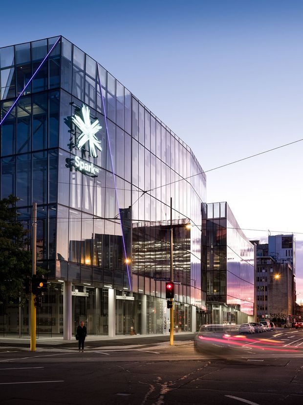 A dusk street view of a large contemporary commercial building with a curved translucent glass facade, extensive dark metal framing, illuminated purple architectural lighting, and prominent Spark branding. The building occupies a prominent urban corner beside a signalised intersection, with surrounding city buildings, streetlights, and light trails from passing vehicles.