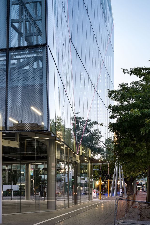 Street-level view of a contemporary urban commercial building with a tall glazed facade, reflective curtain wall, exposed dark structural framing, and perforated metal screens. The building fronts a paved pedestrian streetscape with mature trees and adjacent city buildings visible in reflection. The image emphasizes the exterior architecture and facade detailing.