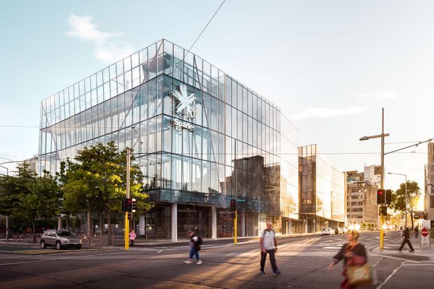 Street-level view of a contemporary multi-storey commercial building on an urban intersection. The building has a transparent glass facade with a large exposed structural grid, glazed corner volumes, and a prominent retail or office sign. Pedestrians, traffic lights, street infrastructure, and neighbouring high-rise buildings reinforce the dense city setting.
