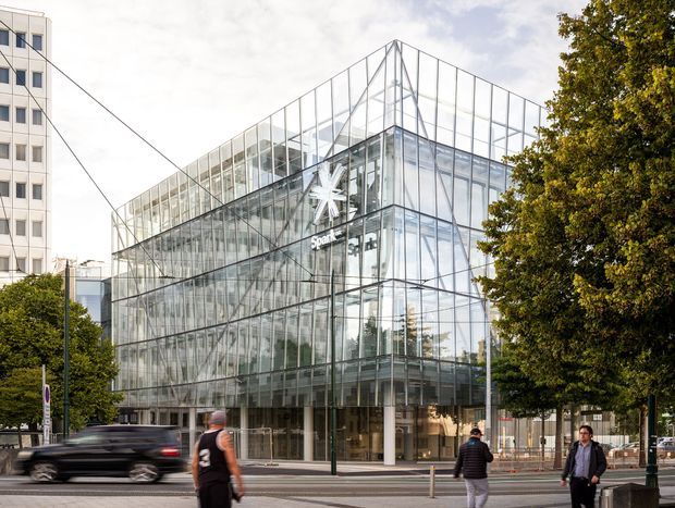 Street-level exterior view of a prominent multi-storey commercial building enclosed in a transparent glass curtain wall and topped with a distinctive angular glazed roof structure. The building occupies an urban streetscape with adjacent office buildings, mature street trees, pedestrians, and passing traffic. A visible Spark logo indicates commercial office or retail use.