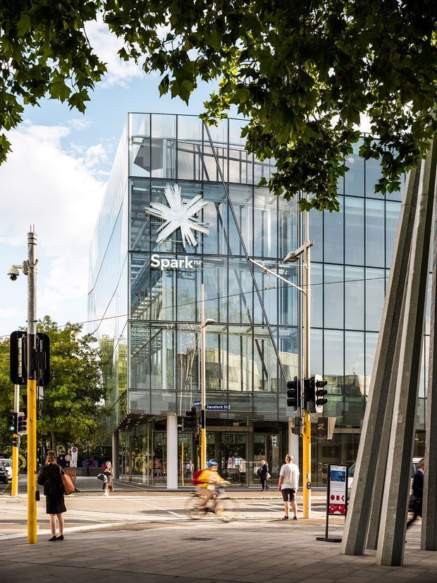 Street-level exterior photograph of a modern multi-storey commercial building with a transparent glass curtain-wall facade and visible internal steel structure. The Spark NZ logo is displayed above the entrance at a busy urban intersection with traffic lights, pedestrians, and cyclists. Large trees frame the upper edge, while substantial angled structural supports are visible in the foreground.