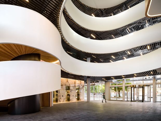A grand multi-level commercial interior lobby or atrium with sweeping curved white balcony slabs and a dramatic central void. Black slatted ceilings, recessed lighting, slender structural columns, expansive glazed entrance doors, and polished grey flooring create a contemporary, high-end architectural setting. A curved timber-lined ceiling feature and dark cylindrical wall element add warmth and contrast near the reception area.