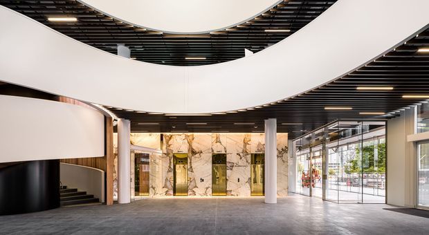 A spacious, sophisticated commercial lobby or entry hall with a dramatic sculptural white curved ceiling installation, black slatted ceiling, polished stone elevator feature wall, cylindrical columns, dark flooring, expansive glass frontage, and a curved black reception or stair enclosure. The space has a high-end contemporary architectural character with strong geometric forms and abundant natural light.