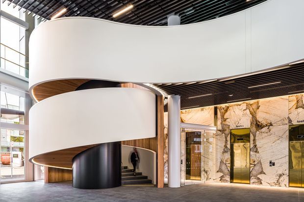 Interior view of a contemporary commercial lobby featuring a dramatic sculptural curved staircase or ramp with smooth white balustrades, dark cylindrical support elements, and warm timber soffits. The space has a black slatted ceiling, polished stone-clad elevator wall with gold-toned lift doors, large glazed openings, and a refined high-end material palette.
