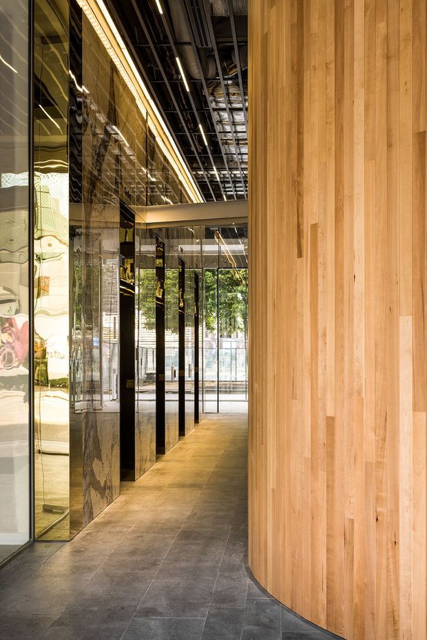 A refined commercial interior corridor or entry space with a dramatic curved wall clad in vertical warm timber boards on the right. The left side features full-height reflective glass and dark polished stone or marble panels, creating layered reflections and a strong sense of depth. Large-format grey floor tiles, linear recessed lighting, and an exposed services ceiling contribute to a contemporary, high-end fit-out. Glazing at the far end looks onto greenery and an outdoor area.