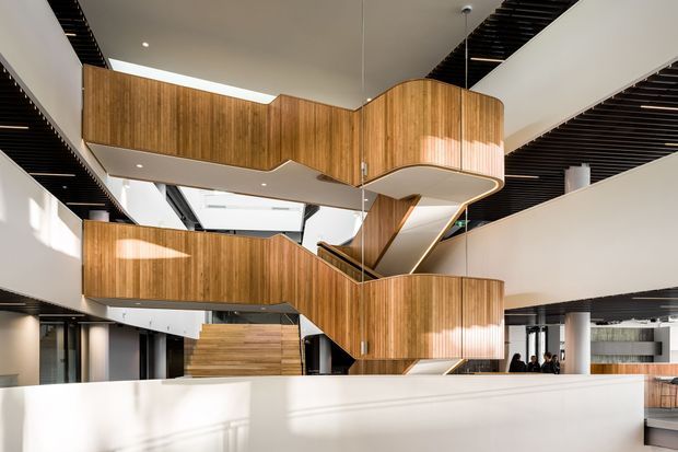 A bright multi-level interior atrium dominated by a sculptural timber-clad stair and circulation structure. Curved vertical-wood balustrades and enclosed stair volumes connect several white floor plates, with exposed black slatted ceiling elements, skylights, and white structural columns. The space has a contemporary commercial or institutional character and strong geometric contrast between warm timber and white surfaces.