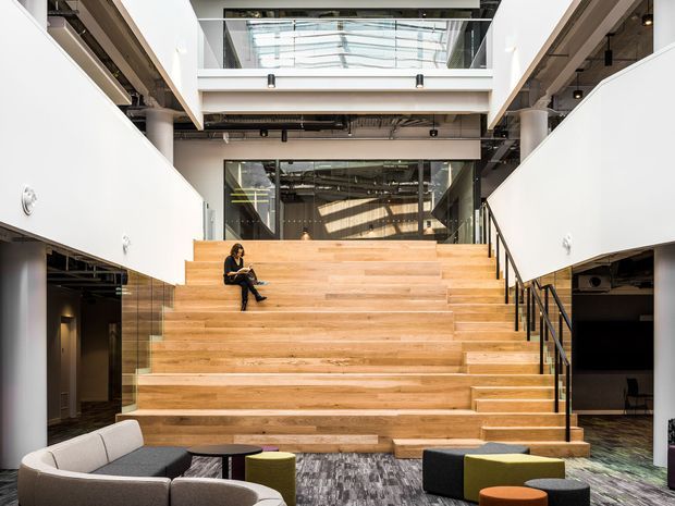 A spacious contemporary commercial-style interior centred on a broad timber stepped stair or tiered seating area. White walls and exposed services frame a double-height atrium with glass balustrades and upper-level glazing. The stair incorporates informal seating, with lounge furniture and small ottomans in the foreground on a patterned carpet. The space has an open, adaptable workplace or public-interior character with strong natural light from overhead glazing.