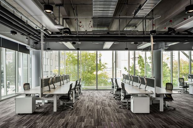 Open-plan contemporary office interior with rows of workstation desks, ergonomic office chairs, large floor-to-ceiling windows, and a fully exposed services ceiling with ducts, cable trays, and suspended lighting. The space uses dark industrial ceiling elements, grey carpet flooring, and neutral workstations, with trees and neighbouring buildings visible outside.