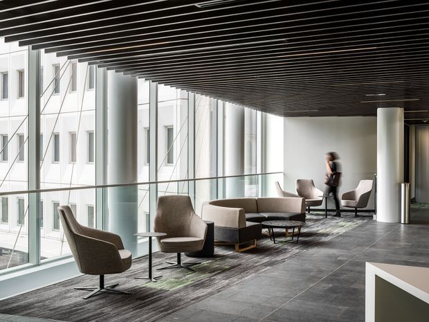 A spacious contemporary commercial lounge or reception area with floor-to-ceiling glazing, a dark linear slatted ceiling, grey tiled flooring, and a refined seating arrangement of upholstered lounge chairs, sofas, and low tables. A glass balustrade overlooks an adjacent building, while cylindrical columns and subtle greenery add structure and softness.