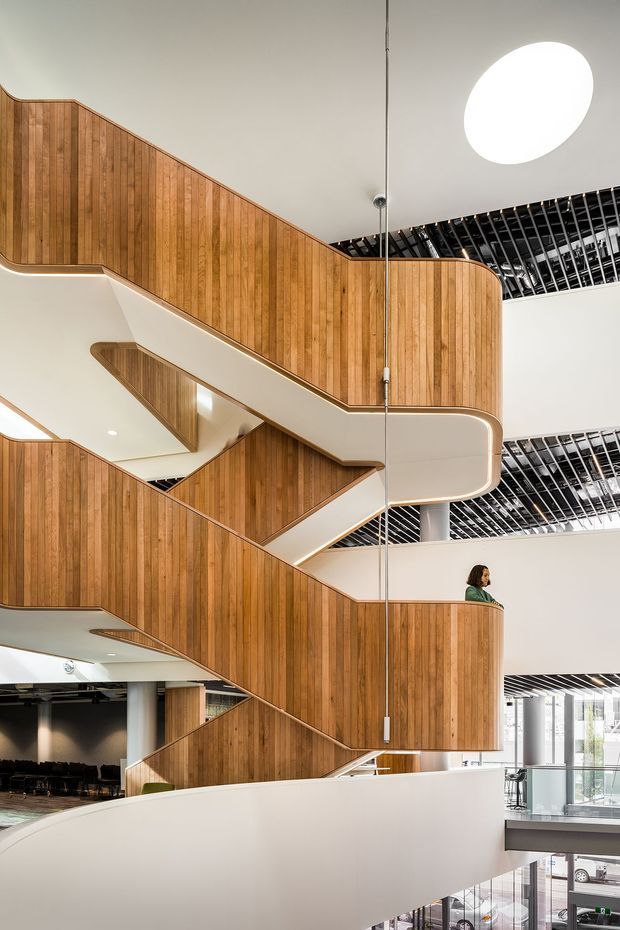Interior architectural photograph of a large multi-level stairway or atrium. A sculptural zigzag staircase with warm vertical timber balustrade cladding and smooth white curved undersides rises through several floors. Black linear ceiling slats, exposed structural elements, glass balustrades, round skylight-like ceiling lighting, and glimpses of an open commercial or institutional interior are visible. The composition emphasizes the dramatic vertical circulation space and double-height volume.