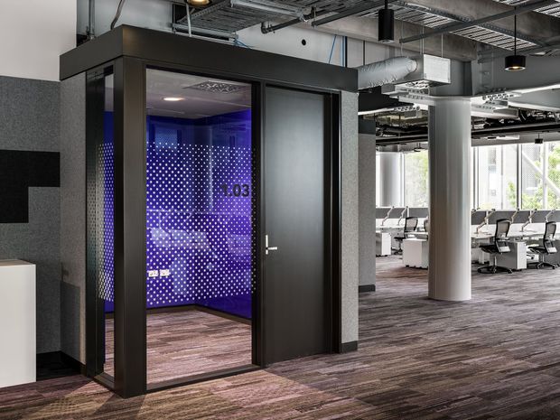 Interior view of a contemporary commercial office with an exposed services ceiling, dark acoustic wall finishes, carpet flooring, and a glazed enclosed room featuring a vivid blue-purple dotted feature wall. Open-plan workstations are visible beyond the enclosure, with cylindrical structural columns and suspended pendant lighting.