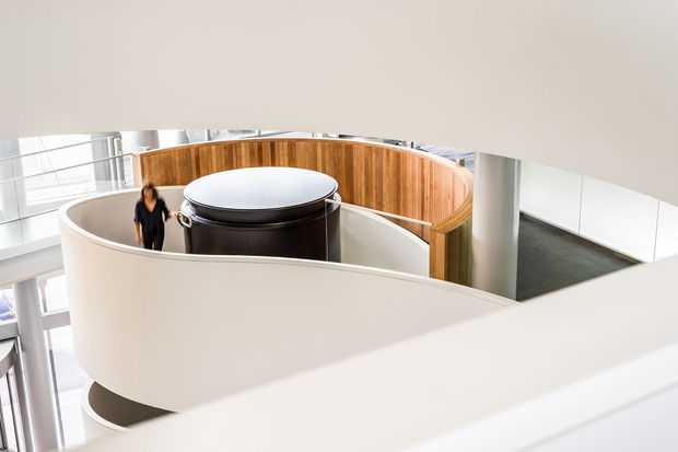 Interior architectural view of a bright, multi-level commercial or public building featuring a sweeping curved white balustrade, a cylindrical dark central structure, curved timber wall panelling, white structural columns, and extensive glazing. The composition emphasizes sculptural circulation and layered mezzanine-like forms.