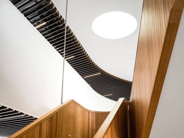 Interior architectural view of a modern stairway or landing, looking upward toward a sweeping curved ceiling void. Warm vertical timber wall panelling and stair balustrades contrast with white sculptural ceiling surfaces, dark exposed linear ceiling battens, recessed lighting, and a large circular skylight or ceiling light. The image emphasizes geometry, material contrast, and dramatic circulation-space design.